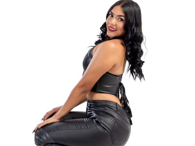 ThaleiaStone, with black curly hair, wearing a black leather crop top and matching pants, sitting and smiling, showing her ass and tight leather pants, with a white background.