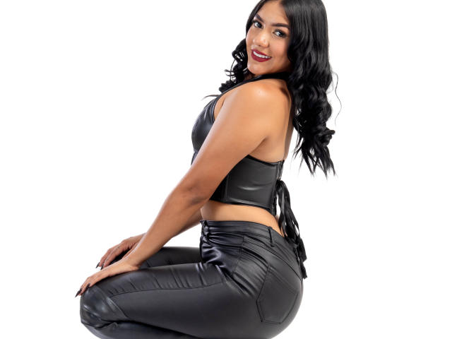 ThaleiaStone with dark skin, long black wavy hair, black leather crop top, black leather pants, kneeling, smiling, showing her ass, medium-sized boobs, red lipstick.