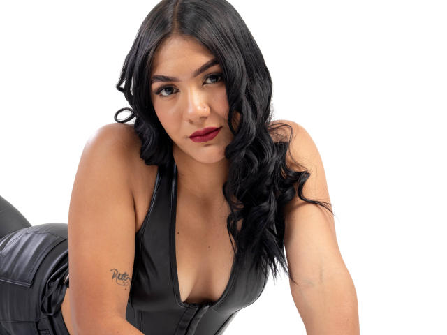ThaleiaStone with black wavy hair, red lipstick, and black leather outfit, leaning forward, showing cleavage, tattoo on left arm, looking at camera, white background.