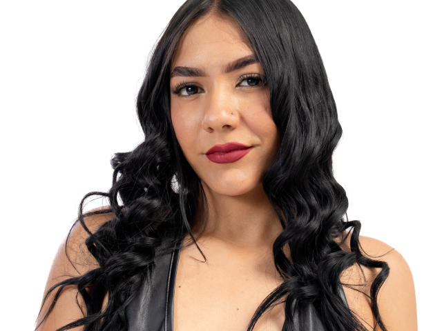 ThaleiaStone has long black wavy hair, red lipstick, and a black sleeveless top, showing a confident, slightly smug expression. Her tits are small and her pussy is not visible.