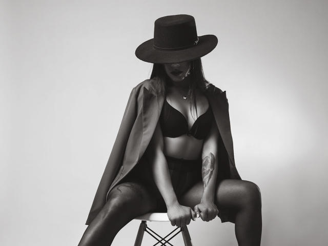 Photo en noir et blanc d'Amberv assise sur un tabouret, portant un chapeau, un soutien-gorge noir et une cape, avec ses seins visibles, des bras tatoués, les jambes écartées, et regardant vers le bas..