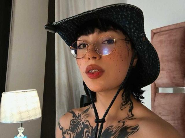 CleoZapata with black hat, round glasses, red lips, freckles, and tattoos on her neck and chest, wearing a strapless top, next to a lit lamp.