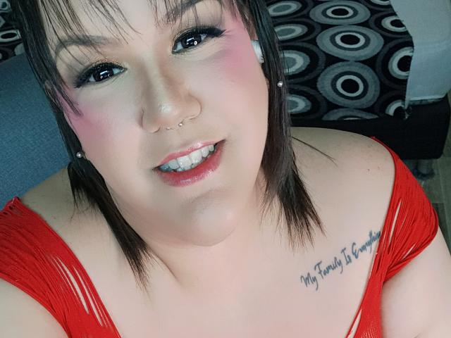 GiiannaMorrys with pink blush, black eyeliner, nose ring, septum piercing, red dress, white earbuds, brown hair, smiling, tattoo on right shoulder, looking at camera.