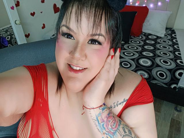 GiiannaMorrys with short black hair, red fishnet top, red nails, nose ring, smile, tattooed arm, holding cheek, black cat ears, pink eye makeup, red bracelet, and rose tattoo.