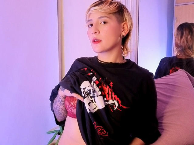 DannyRouds, avec ses courts cheveux blonds, portant un t-shirt noir avec un motif, laissant entrevoir des sous-vêtements en dentelle rouge et des bras tatoués, assise sur un coussin rose, regardant l'appareil photo..