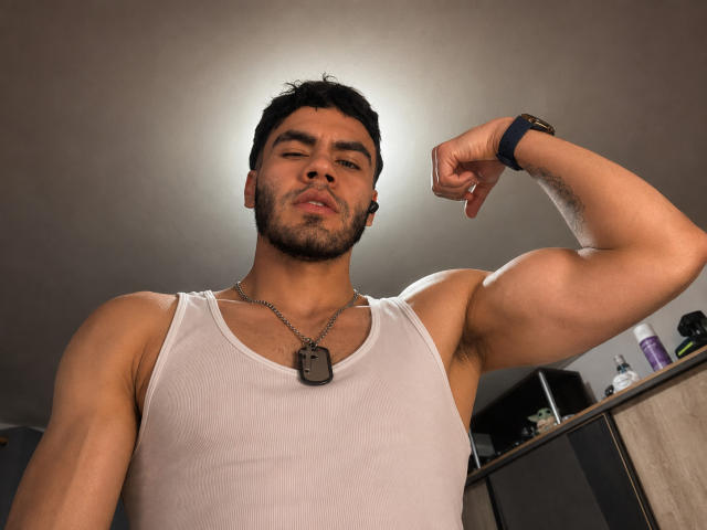 NastyX69, musculoso, exibindo o braço, vestindo uma camiseta branca, usando pulseiras de identificação, um relógio preto, cabelo curto e escuro, barba, pele clara, olhando para baixo, expressão confiante, ambiente interno, foto tirada de baixo..