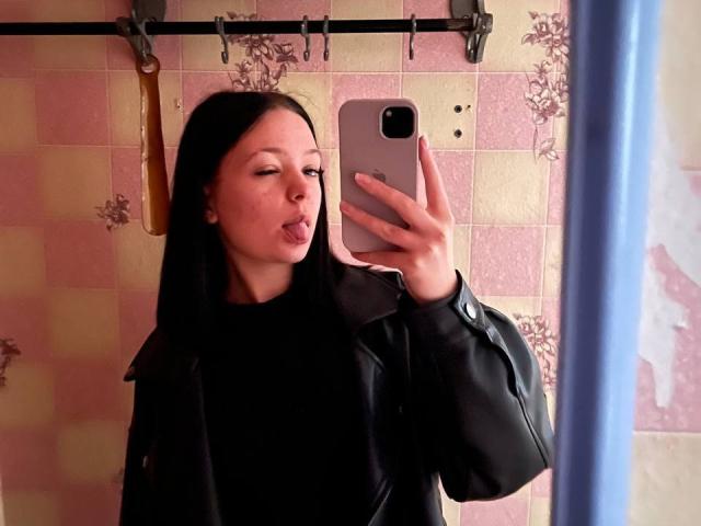 JestineBaumann with black hair, black shirt, and black leather jacket, taking a mirror selfie with tongue out, holding a silver phone, in a tiled bathroom.