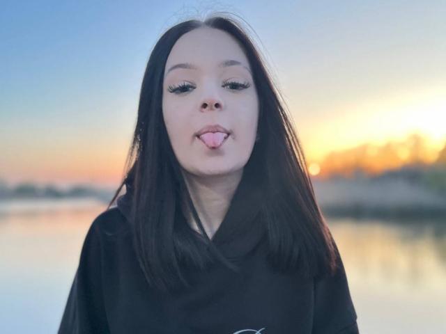 JestineBaumann has straight dark brown hair, fair skin, and is sticking out her tongue. She's wearing a black hoodie with long sleeves. Her eyes are half-open.