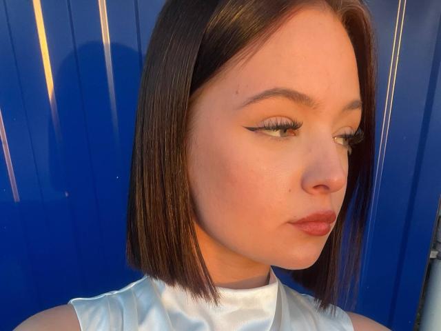 JestineBaumann has a short brown bob, white eyeliner, and is wearing a shiny white top. Her face is slightly turned to the side, showing a calm expression.