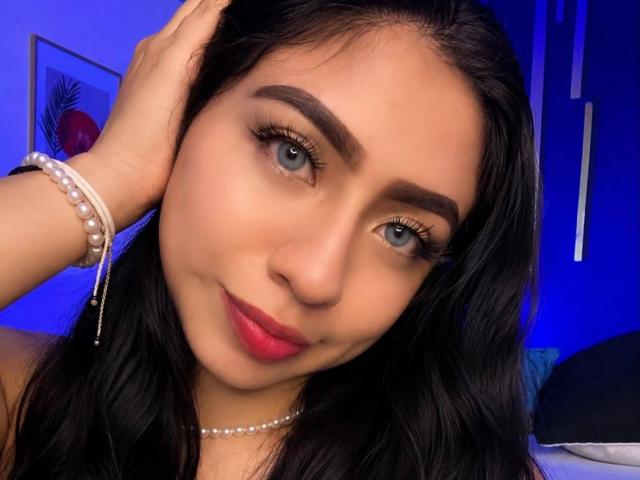 ZoeRedds with blue eyes, dark eyebrows, red lipstick, black hair, and a pearl bracelet, touching her head with a slight smile, looking into the camera.
