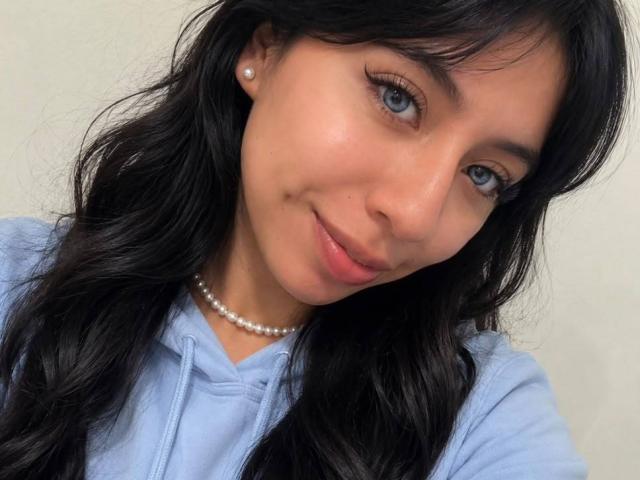 ZoeRedds has long black hair, blue eyes, and a slight smile with a pearl necklace, wearing a light blue hoodie, and a small beauty mark on her right cheek.