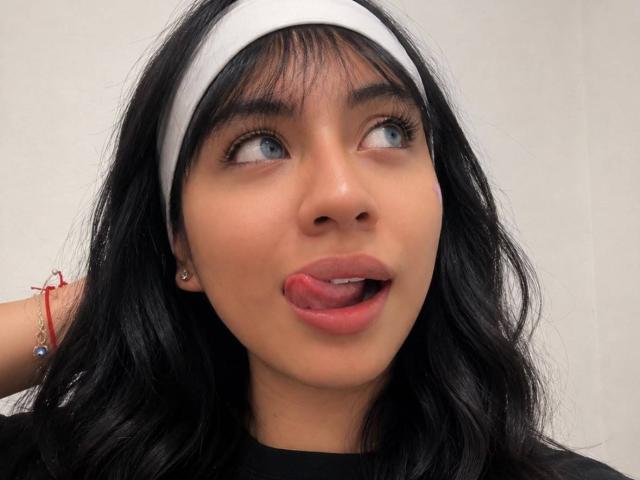 ZoeRedds has black wavy hair, blue eyes, and a white headband. She's sticking her tongue out, looking to the side with a playful expression. She's wearing a black top.