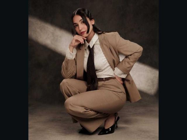MindyLove squatting in a beige suit, white shirt, brown tie, and black heels, with a hand near her face and a subtle smile. Her ass is small and cute.