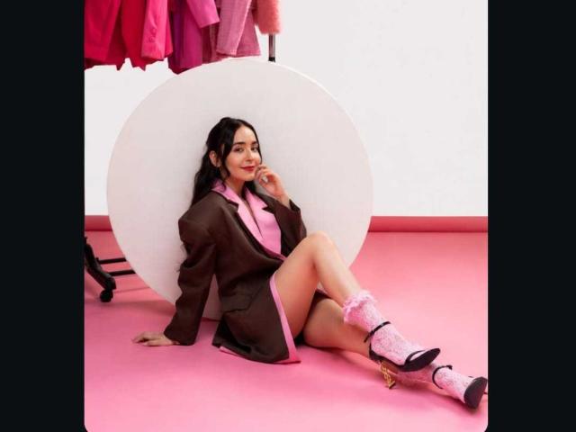 MindyLove with black hair, brown jacket with pink trim, pink socks, and black heels, leaning against white circle, pink floor, and pink clothes hanging in background.