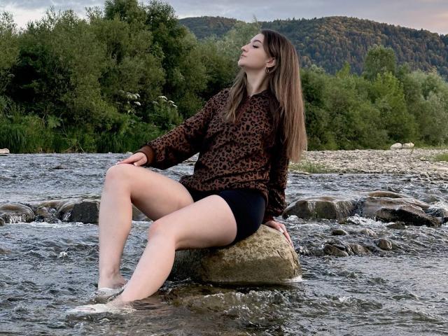 CyrenaLuxe with long brown hair, leopard print sweater, and black shorts, sits on a rock in a flowing river, legs in water, eyes closed, looking up.