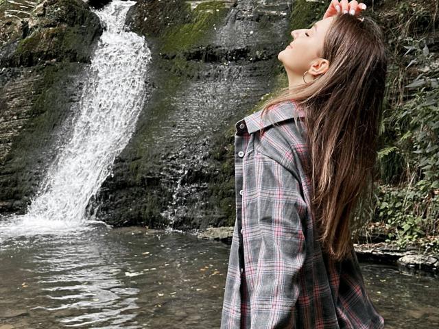 CyrenaLuxe with long brown hair, eyes closed, head tilted back, wearing an oversized plaid shirt, stands by a waterfall, her hand touching her head, water splashing below.