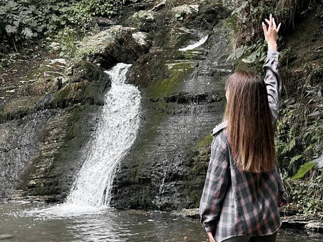 CyrenaLuxe with long brown hair, plaid shirt, and shoulder bag, stands with arm raised, facing small waterfall. Her back is to the camera, water splashes below.