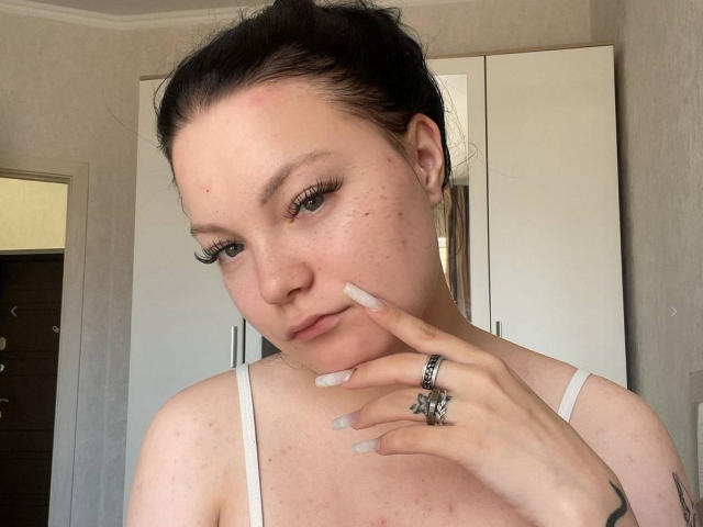 Peg Huntzinger tem pele com sardas, cabelo escuro preso em um coque, cílios longos, uma camiseta branca, anéis e uma tatuagem na mão direita, que se estende até o queixo..