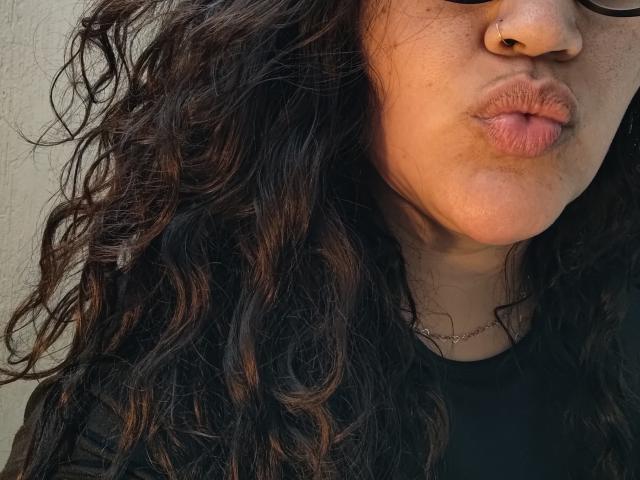 Brendabbew's puffy brown lips, nose ring, curly dark brown hair, wearing black top, visible necklace, partially obscured glasses, close-up shot, tan skin.