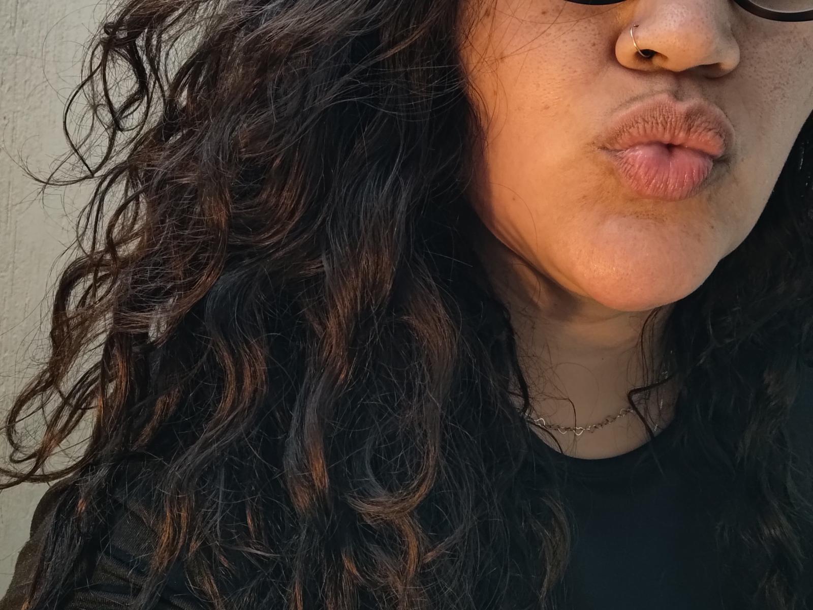 Brendabbew's puffy brown lips, nose ring, curly dark brown hair, wearing black top, visible necklace, partially obscured glasses, close-up shot, tan skin.