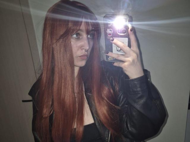 RayBeliles, con cabello rojo largo, chaqueta de cuero negro, sosteniendo un teléfono en un selfie frente al espejo, con el flash en la cara, un piercing en la nariz y esmalte de uñas oscuro, mostrando sus pechos y glúteos..