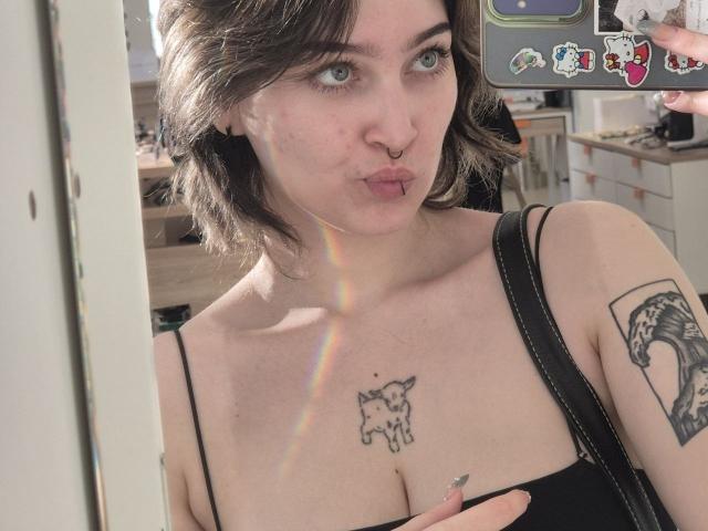 RayBeliles with short brown hair, nose ring, and tattooed chest, pouting lips, rainbow reflection, black strap top, holding phone with stickers, visible tattoo on right arm.
