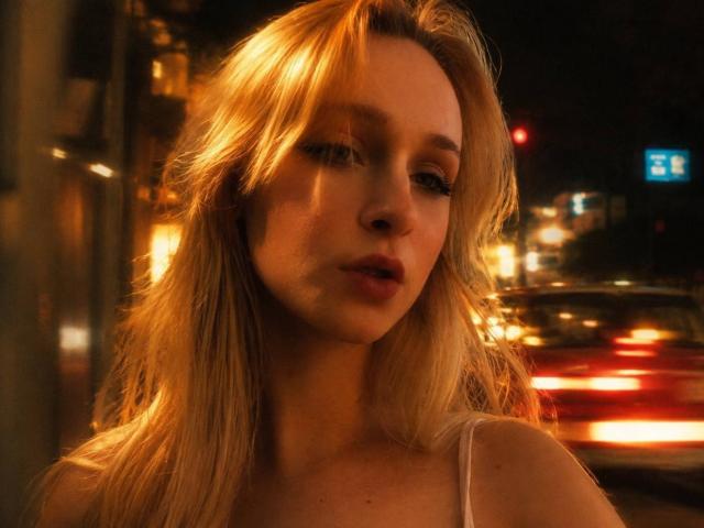 FlyFreedom has blonde hair, full lips, and a slight pout. Her eyes are half-closed, and she's wearing a white top, with soft, warm light highlighting her face.