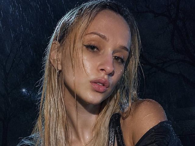 FlyFreedom's wet, blonde hair clings to her face, glistening with raindrops. She has pouty, pink lips and a slightly melancholic expression. Her bare shoulders are visible.