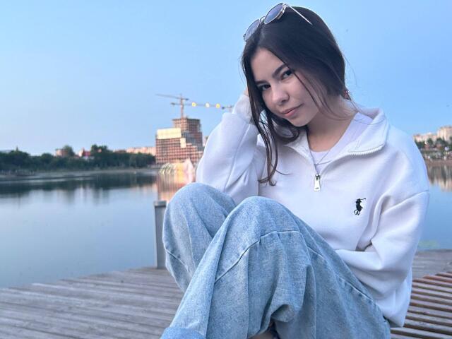 EmeraldCrivaro sitting on a wooden dock, wearing a white hoodie with a small black logo, blue jeans, and sunglasses on her head, smiling with her hand in her hair.