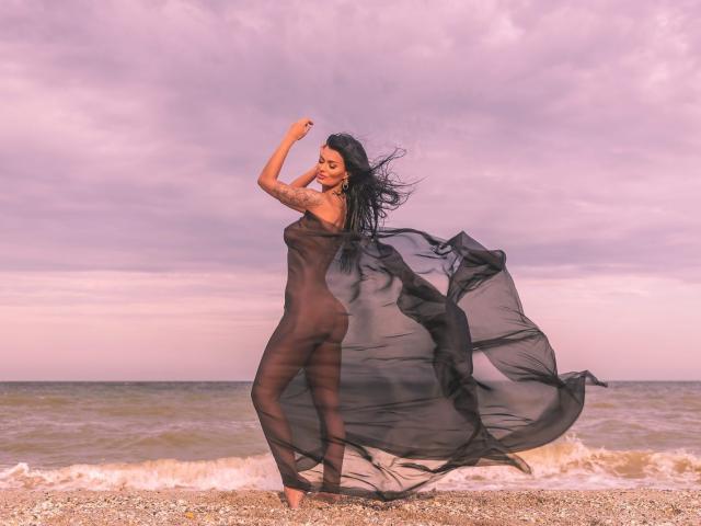 Amy Christie, con cabello negro largo, vestida con un vestido negro transparente, de pie en una playa pedregosa, con su vestido ondeando, mostrando sus pechos y glúteos, bajo un cielo rosa-morado..