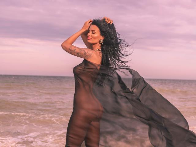Amy Christie, con lunghi capelli neri, indossa un abito nero trasparente che rivela i suoi genitali e il sedere, è in piedi vicino all'oceano, con una mano tra i capelli, ha tatuaggi sul braccio, il cielo è rosa..