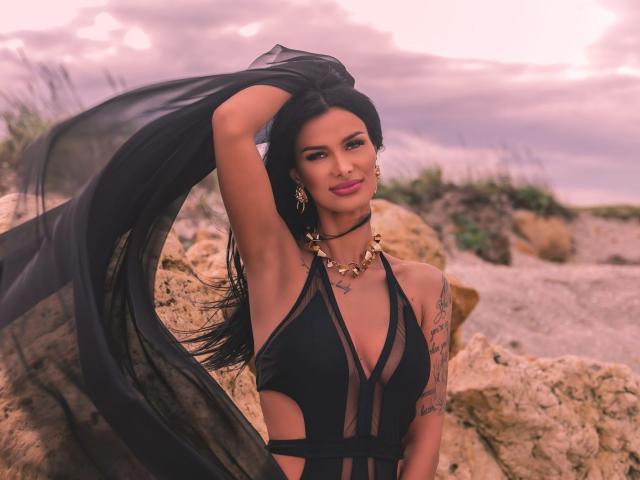 AmyChristie with long black hair, wearing a sheer black dress, exposing her tits, gold necklace, tattoos on her arm, standing by rocks, holding her hair, pink sunset sky.