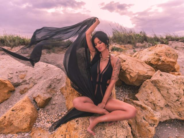 AmyChristie with black sheer dress, holding fabric above her head, sitting on rocks, legs crossed, tattoos on left arm, gold necklace, purple sunset, wind blowing through her hair.