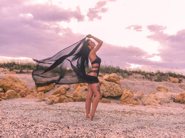 AmyChristie with long black hair, wearing a black sheer dress, standing on pebbles, arms raised, dress flowing, showing her ass and tattoo on right thigh, under a pink sky.