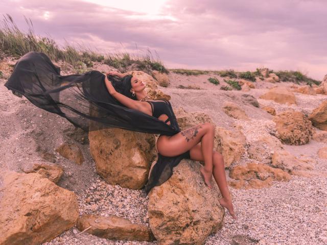 Amy Christie mit blonden Haaren, einem schwarzen, transparenten Kleid und Tattoos, liegt auf einem felsigen Strand, die Beine gekreuzt, das Kleid flattert im Wind, am Horizont ein rosafarbener Himmel, ihr Hintern und ihre Brüste sind teilweise sichtbar..