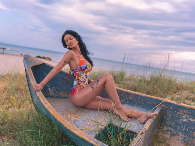 AmyChristie with tan skin and black hair, sitting in a worn-out blue boat, wearing a colorful, tight, one-piece swimsuit, showing her tits and ass, with tattoos on her thigh.
