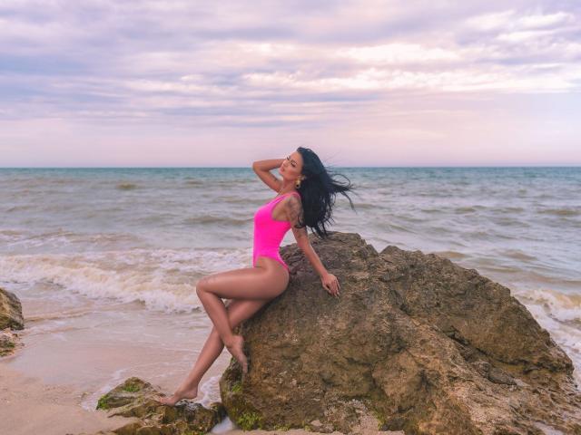 AmyChristie with dark hair, pink one-piece, sits on rock, hand in hair, legs crossed, ocean waves, sunset sky, her ass and tits highlighted, beach, rocks, water.