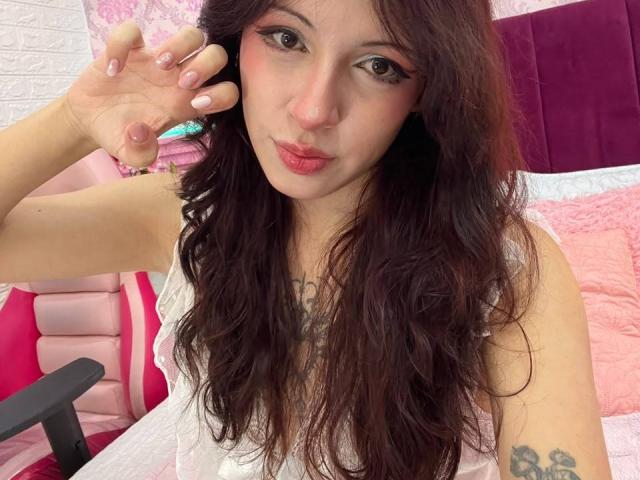 Valery Beaumont mit langen, braunen Haaren, pinkem Eyeliner und pinkem Lippenstift, die eine Hand mit pinken Nägeln hochhält, trägt ein weißes Oberteil und zeigt ein Tattoo auf ihrem linken Arm..