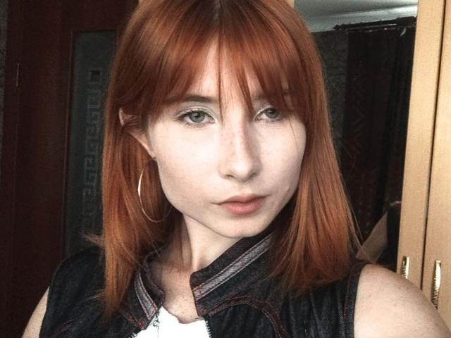 KasandraGrothe has red hair, green eyes, pale skin, and wears a black sleeveless top with a white shirt underneath. She's looking at the camera with a neutral expression.