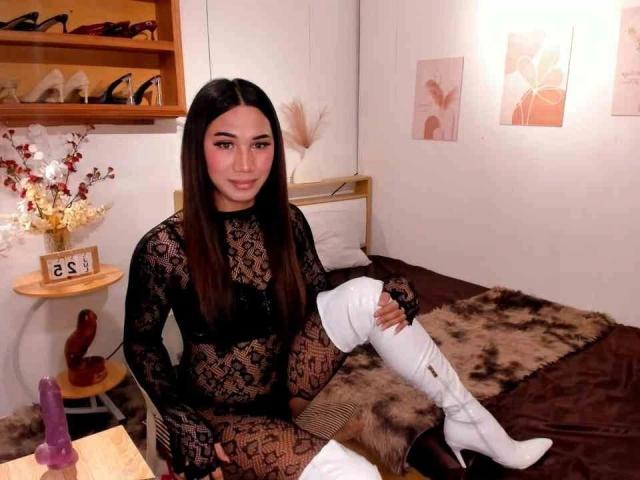 Matilda Santos – attraktiv mit langen, braunen Haaren, trägt einen schwarzen Spitzen-Body und weiße, hohe Stiefel, sitzt auf einem braunen Bett mit einer Felldecke, die Beine gekreuzt, sexy, Zimmer mit einem Holzregal..