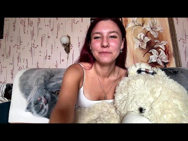 CristieRazey, con cabello rojo, camiseta blanca, sosteniendo un osito de peluche blanco y un cojín con la cara de un gato gris, mirando directamente a la cámara..