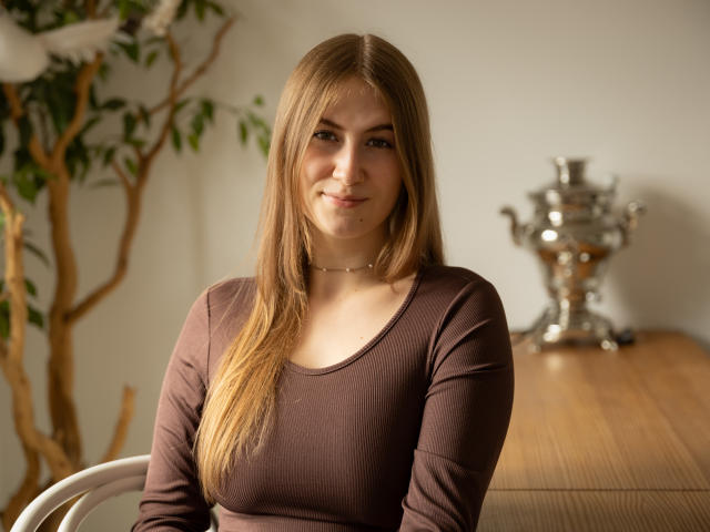 MeganCandy mit langen blonden Haaren, einem braunen Langarmshirt und einer Halskette, lächelt sanft und zeigt kleine Brüste. Sie sitzt vor einem Holztisch mit einer silbernen Vase..