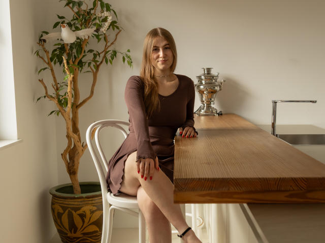 MeganCandy, com cabelos castanhos longos, vestido marrom, sentada em uma cadeira branca, pernas cruzadas, unhas vermelhas, mesa de madeira, vaso de prata, planta em vaso, sorrindo, pele clara, luz suave..