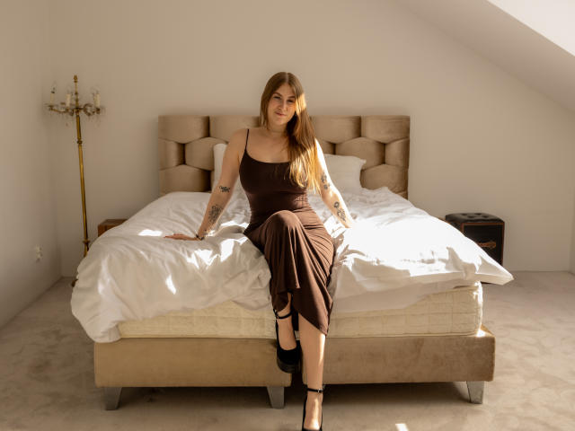 MeganCandy, met lang bruin haar, gekleed in een bruine jurk en zwarte hakken, zit op een bed met witte lakens en laat haar tatoeages en haar achterwerk zien. Slaapkamer met een messing wandlamp..