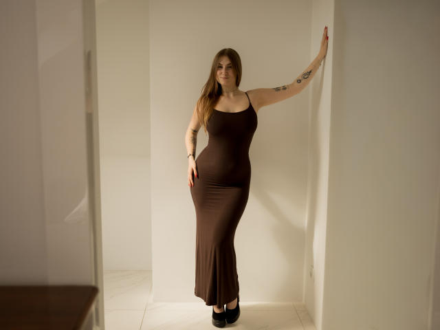 MeganCandy, de pie en un pasillo blanco, vestida con un vestido marrón ajustado que resalta sus curvas, con tatuajes en su brazo derecho, tacones negros y cabello largo y castaño..