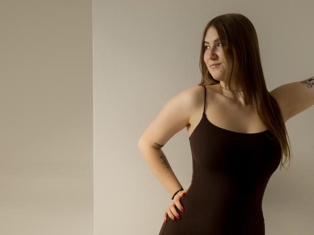 MeganCandy, com cabelos castanhos longos, vestindo um vestido preto com alças finas, em pé com a mão na cintura, mostrando tatuagens e um sorriso discreto, em frente a uma parede branca lisa..