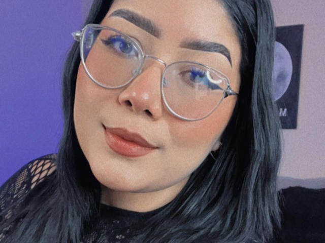 AlexaKiim with blue eyes, wearing clear glasses, black lace top, and black hair, has a cute face and subtle smile. Her tits are not visible in this shot.