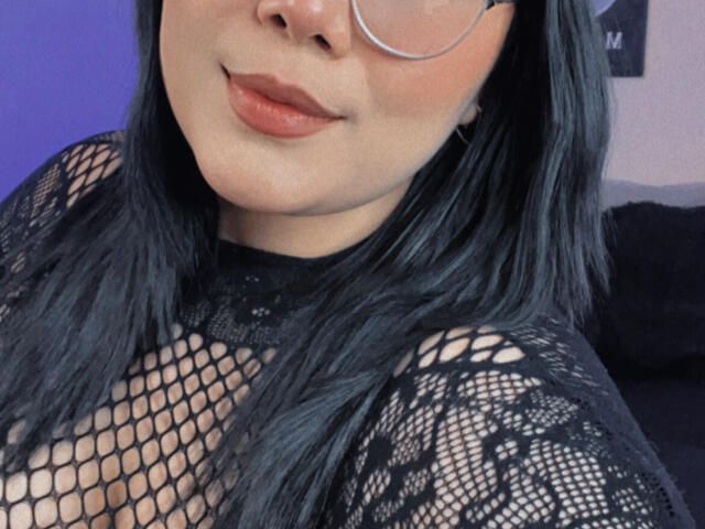 AlexaKiim with black fishnet top, showing her tits through the net, wearing glasses, black hair, and pink lipstick, smiling slightly. Her skin is light brown.