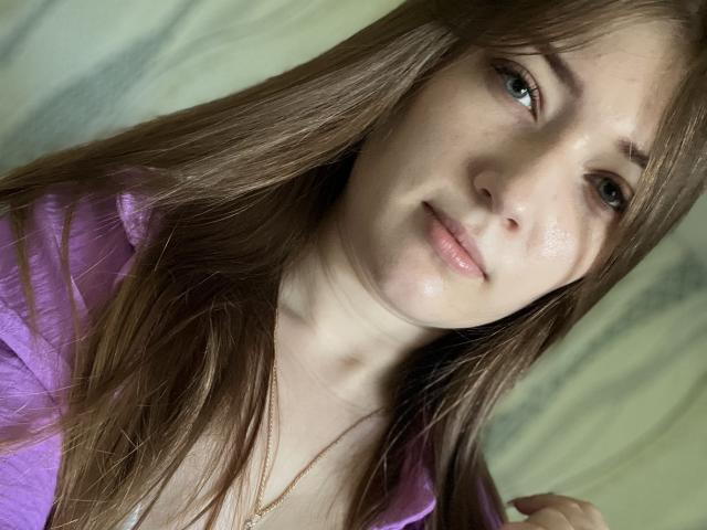 Krrissty has light brown hair, green eyes, and a subtle smile. She's wearing a purple shirt and a gold necklace, with a slightly shiny, smooth face.