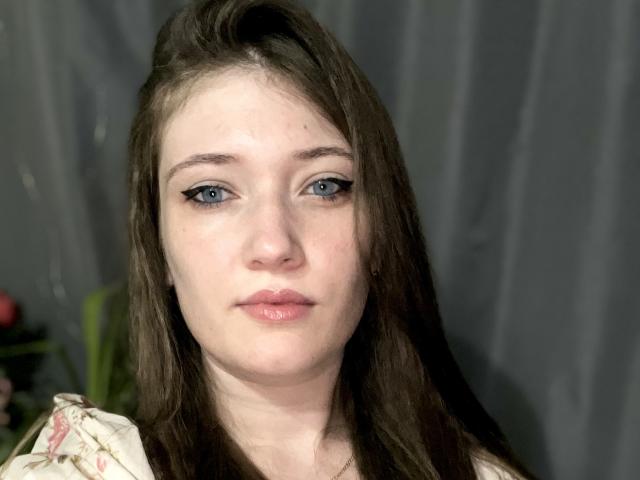 Krrissty has blue eyes, light skin, brown hair, and pinkish lips. She's wearing a floral top, looking straight at the camera with a neutral expression.